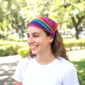 Op de afbeelding: Een kleurrijke regenboog gestreepte hoofdband. De hoofdband heeft een levendige reeks kleuren, waaronder roze, rood, oranje, geel, groen, blauw en paars. De hoofdband is aan de achterkant vastgebonden. De persoon draagt een wit t-shirt.