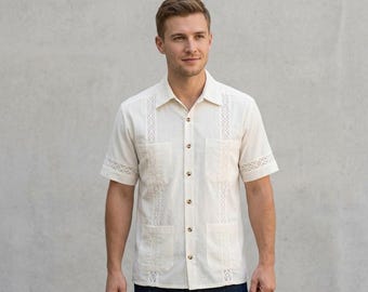Può includere: Una camicia a maniche corte color crema con pannelli verticali di ricamo in pizzo bianco, colletto classico e quattro tasche frontali. La camicia è realizzata in tessuto testurizzato e presenta bottoni di colore marrone.