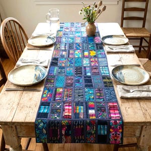 May include: A rectangular table runner featuring a patchwork design of various colorful woven fabric strips in shades of blue, pink, yellow, and green, arranged in a grid pattern, centered on a wooden dining table with plates and silverware.