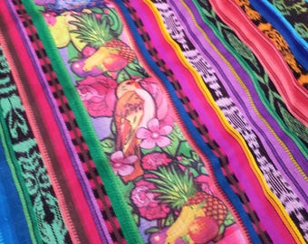 Puede incluir: Un tejido vibrante con rayas verticales en tonos rosa, morado, azul, verde, amarillo y negro. Un panel central muestra un patrón repetitivo de frutas tropicales coloridas, incluyendo piñas, y aves rodeadas de flores de color rosa.