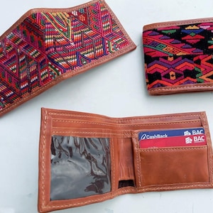 May include: Three leather wallets. Two wallets feature colorful woven fabric designs in red, pink, and green. The third wallet is open, displaying card slots and a clear ID window. The open wallet is brown.