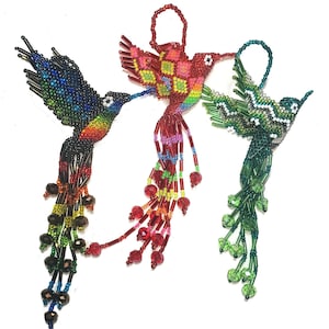 Op de afbeelding: Drie kleurrijke, met kralen versierde kolibri-ornamenten. Eén is voornamelijk blauw met regenboogaccenten, een andere rood met een dambordpatroon en de derde groen. Elk heeft een lus om op te hangen en een kralenstaart.