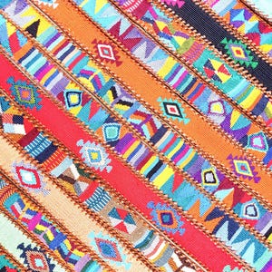 Puede incluir: Textil colorido con un patrón de rayas diagonales. El diseño presenta formas y patrones geométricos en tonos rojos, naranjas, azules, amarillos y morados. Los bordes están cosidos con hilo marrón. Diseño vibrante.