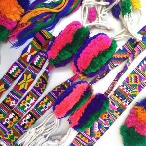 May include: Vibrant, handwoven textile belts with colorful geometric patterns and fluffy pom-poms. The belts feature a variety of colors, including purple, green, pink, yellow, and blue. These decorative belts are perfect for adding a pop of color to any outfit.