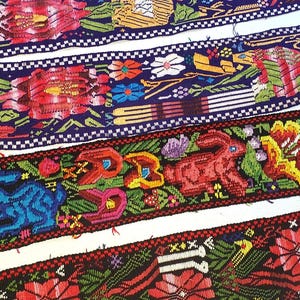 May include: Close-up of colorful woven textiles with intricate patterns. The designs feature floral and animal motifs, including rabbits and birds, in vibrant hues of red, blue, yellow, and green. The textiles have white and dark blue borders.