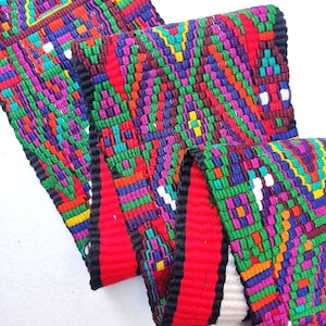 May include: A colorful woven textile with a geometric pattern. The design features a vibrant mix of red, green, purple, blue, and yellow. The edges are trimmed with black and red fabric. The textile is folded, showcasing its intricate details.