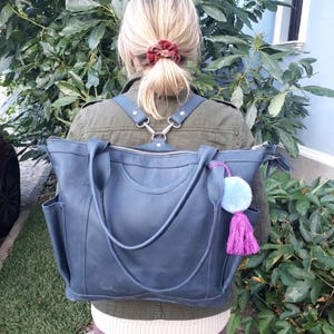 Könnte beinhalten: Eine große Tragetasche aus marineblauem Leder mit zwei Tragegriffen und verstellbaren Rucksackgurten. Die Tasche verfügt über einen Reißverschluss oben, Seitentaschen und einen hellblauen Pompon mit einer leuchtend pinken Quaste an der Seite.