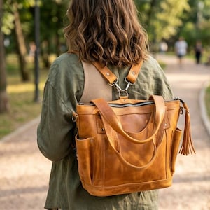 Puede incluir: Una mochila y bolso de mano convertible de cuero color canela con cierre de cremallera superior, dos bolsillos laterales y una borla de cuero larga en el lateral. El bolso incluye correas ajustables conectadas mediante clips de metal.