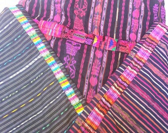 Puede incluir: Primer plano de textiles tejidos coloridos. Las telas presentan un fondo oscuro con rayas verticales en blanco, rosa y morado. Los bordes están adornados con un borde vibrante de colores del arcoíris. Los textiles están doblados y en capas, mostrando los intrincados patrones.
