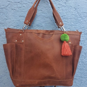 May include: A brown leather tote bag with two exterior pockets and a zipper closure. The bag has two handles and a detachable shoulder strap. A green pom-pom and orange tassel hang from the zipper. The bag is against a blue wall.