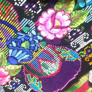 以下が含まれることがあります： 黒い布地に施された緻密でカラフルな刺繍。鮮やかなピンクのバラと緑の葉、明るい青の花のモチーフ、そして黄色、赤、緑の糸でクロスステッチ風に縫い付けられた幾何学模様が特徴です。