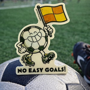 May include: A soccer ball sticker with a cartoon soccer ball character holding a referee flag. The sticker has the text "NO EASY GOALS!". The background shows a soccer field and a pair of cleats.