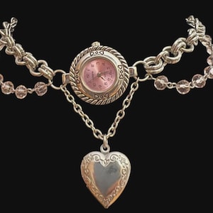May include: Silver-toned watch necklace with a pink watch face and a heart-shaped pendant. The watch is connected to a chain and beaded strands. The watch face has small heart-shaped markers.