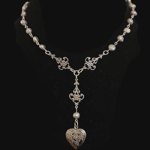 Puede incluir: Collar plateado con un colgante en forma de corazón. El collar presenta una delicada cadena con pequeñas cuentas redondas y detalles florales de filigrana. El relicario está grabado con un diseño de rosas.