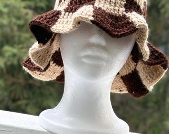 May include: A handmade crochet bucket hat in a brown and beige checkerboard pattern. The hat has a wide, ruffled brim and is displayed on a white mannequin head. The background is a blurred view of green foliage.