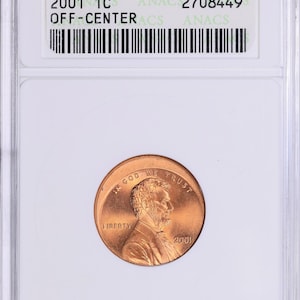 May include: A copper-colored 2001 1C penny graded MS 65 RED by ANACS, with an off-center strike. The coin is encased in a clear plastic holder. The obverse features a portrait and the word "LIBERTY".