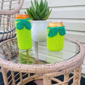 K&ouml;nnte beinhalten: Zwei limettengr&uuml;ne H&auml;kel-Dosenk&uuml;hler mit gr&uuml;nen Bl&auml;ttern sitzen auf einem Tisch mit Glasplatte. Eine Aloe Vera-Pflanze in einem wei&szlig;en Topf befindet sich im Hintergrund. Ein Korbstuhl ist teilweise sichtbar.