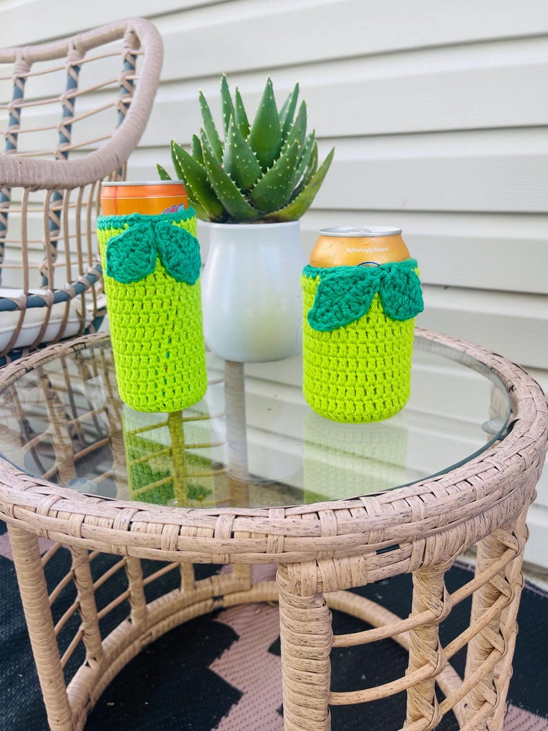 K&ouml;nnte beinhalten: Zwei limettengr&uuml;ne H&auml;kel-Dosenk&uuml;hler mit gr&uuml;nen Bl&auml;ttern sitzen auf einem Tisch mit Glasplatte. Eine Aloe Vera-Pflanze in einem wei&szlig;en Topf befindet sich im Hintergrund. Ein Korbstuhl ist teilweise sichtbar.