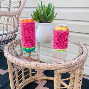 K&ouml;nnte beinhalten: Zwei pinke geh&auml;kelte Getr&auml;nkedosen-K&uuml;hler mit Wassermelonen-Design stehen auf einem Tisch mit Glasplatte. Die K&uuml;hler haben gr&uuml;ne und wei&szlig;e Akzente. Eine Topf-Aloe-Pflanze und ein Korbstuhl sind im Hintergrund.