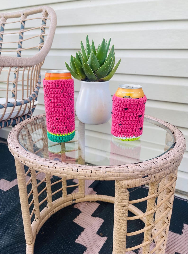 K&ouml;nnte beinhalten: Zwei pinke geh&auml;kelte Getr&auml;nkedosen-K&uuml;hler mit Wassermelonen-Design stehen auf einem Tisch mit Glasplatte. Die K&uuml;hler haben gr&uuml;ne und wei&szlig;e Akzente. Eine Topf-Aloe-Pflanze und ein Korbstuhl sind im Hintergrund.