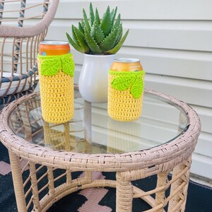 K&ouml;nnte beinhalten: Zwei gelbe H&auml;kel-Dosenk&uuml;hler mit limettengr&uuml;nen Bl&auml;ttern sitzen auf einem Glastisch aus Korbgeflecht. Eine Topf-Aloe-Pflanze und ein Korbstuhl befinden sich im Hintergrund. Die K&uuml;hler sind daf&uuml;r gemacht, Getr&auml;nke kalt zu halten.