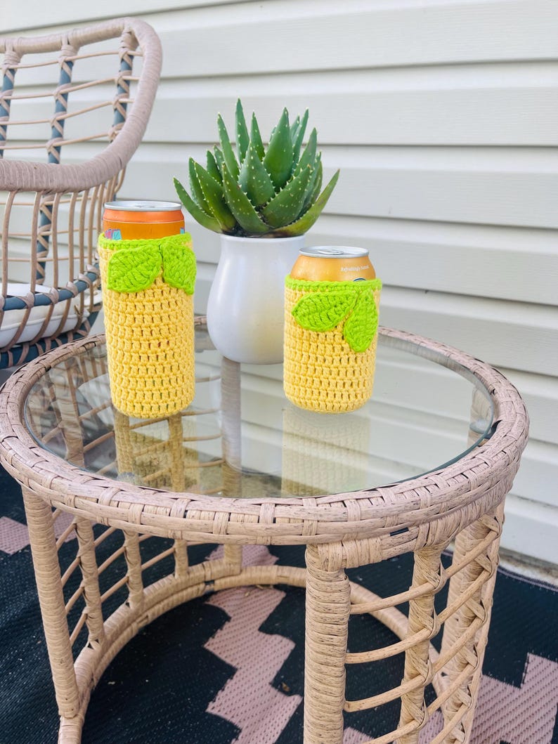 K&ouml;nnte beinhalten: Zwei gelbe H&auml;kel-Dosenk&uuml;hler mit limettengr&uuml;nen Bl&auml;ttern sitzen auf einem Glastisch aus Korbgeflecht. Eine Topf-Aloe-Pflanze und ein Korbstuhl befinden sich im Hintergrund. Die K&uuml;hler sind daf&uuml;r gemacht, Getr&auml;nke kalt zu halten.