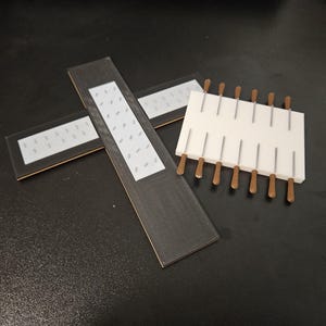 以下が含まれることがあります： 暗い表面にある3つの長方形のオブジェクトの俯瞰図。2つは長く、粘着ストリップが付いた濃い灰色です。3つ目は、小さなナイフが挿入された白いブロックで、それぞれに茶色のハンドルと銀色の刃が付いています。