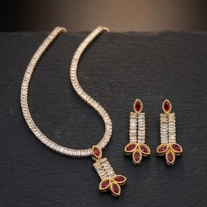 以下が含まれることがあります： ゴールドトーンのネックレスとマッチするイヤリングのセット。ネックレスとイヤリングは、クリアと赤の宝石を使用した線形デザインです。ネックレスには赤い宝石のクラスターが付いたペンダントが付いています。イヤリングは垂直デザインです。