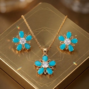 May include: A gold-toned jewelry set featuring a necklace and matching earrings. Each piece showcases a flower design with turquoise-colored petals and a central clear gemstone. The necklace pendant and earrings are displayed on a gold-colored surface.
