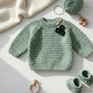 Op de afbeelding: Een lichtgroene, handgehaakte baby trui met een textuursteek, houten knopen en zwarte bloemversieringen. Bijpassende babyslofjes en een houten bijtring zijn ook aanwezig. Een bol wol en een zachte deken maken de scène compleet.