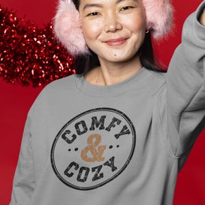 May include: A gray sweatshirt with the words "COMFY & COZY" in a distressed black and brown design. The person in the image is wearing pink earmuffs. The background is red.