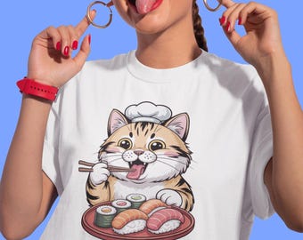 May include: White t-shirt featuring a cartoon cat chef enjoying sushi. The cat, wearing a chef's hat, holds chopsticks and is seated in front of a plate of sushi rolls and nigiri. The image is set against a blue background.