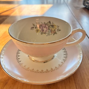 Peut inclure: Service à thé vintage composé d'une tasse rose pâle avec un bord doré et un motif floral, posée sur une soucoupe assortie. La soucoupe présente une bordure décorative dorée et blanche. L'ensemble est posé sur une table en bois.