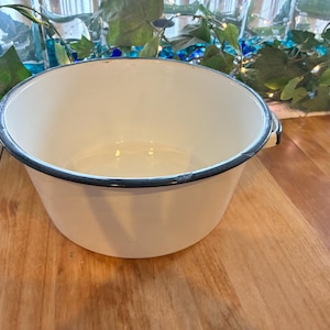 May include: A white enamelware bowl with a dark blue rim and handles. The bowl is sitting on a wooden surface. The bowl is round and has a wide opening.