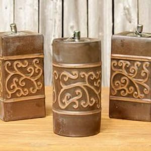 May include: Three brown decorative candle holders with a scroll design. The candle holders are rectangular and cylindrical, with a metal top. The scroll design is a lighter brown color than the base. The candle holders are on a wooden surface.