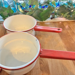 May include: Two white enamel saucepans with red handles and rims. The pans are sitting on a wooden surface. The handles have a hole at the end for hanging. The pans are empty.