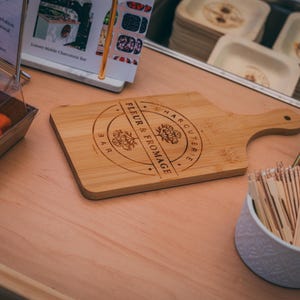 Puede incluir: Una tabla de cortar de madera clara con un asa, grabada con "Fleur & Fromage Bar". La tabla está sobre una superficie marrón claro. También se ve un recipiente con palillos y un recipiente con comida.