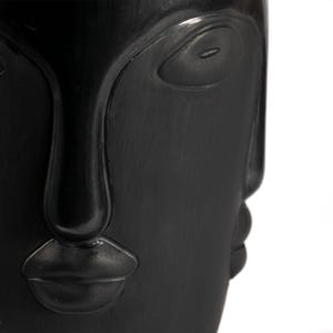 May include: A close-up of a black decorative face sculpture. The sculpture features a stylized face with prominent features, including a large eye, a long nose, and full lips. The surface has a textured appearance.