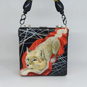 May include: A square handbag with a yellow handle and black accents. The bag features a painting of a light-colored fox on a red background against a black backdrop with white lines. The handle is arched.
