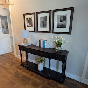 May include: A black console table with three drawers and a lower shelf, adorned with framed black and white photographs, a lamp, books, and vases of flowers. The table is set against a neutral wall with a wooden floor.