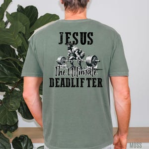 May include: Sage green t-shirt with a graphic on the back. The graphic features an illustration of a weightlifter with the text "JESUS The Ultimate DEADLIFTER" in black. The shirt is a solid color.