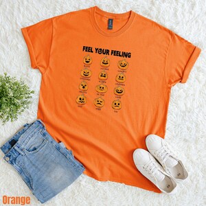 May include: Orange t-shirt with the text "FEEL YOUR FEELING" and pumpkin faces representing different emotions. The shirt is paired with blue jeans and white sneakers. A small green plant is visible.