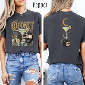 May include: A charcoal gray t-shirt featuring a graphic print of a margarita glass, books, candles, and a crescent moon. The front design includes the text "Put the lime in the COCONUT" and "AND DRINK IT ALL UP". The back features a smaller matching graphic.