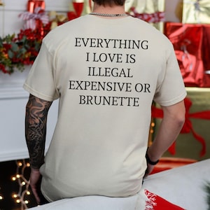May include: A beige t-shirt with the text "EVERYTHING I LOVE IS ILLEGAL EXPENSIVE OR BRUNETTE" printed in black. The shirt is short-sleeved and has a crew neck. The person wearing the shirt has a tattoo on their left arm.