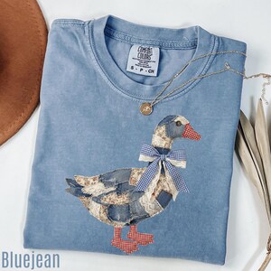 May include: A blue Comfort Colors t-shirt with a patchwork duck design. The duck features denim, floral, and lace fabrics, a gingham bow, and red checkered feet. A gold necklace with a pendant is draped on the shirt. The word "Bluejean" is at the bottom.