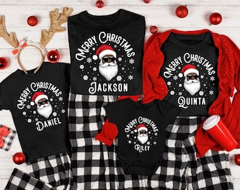 May include: A family Christmas pajama set with black tops and a onesie, each displaying a Santa Claus graphic, paired with black and white checked pajama bottoms. The shirts are personalised with names: Daniel, Jackson, Quinta, and Riley. Red baubles are scattered around.