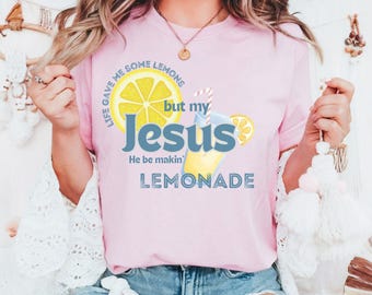 May include: Pink t-shirt with the text "Life gave me some lemons but my Jesus He be makin' lemonade" and a graphic of a lemon and a glass of lemonade. The shirt is a light pink color.