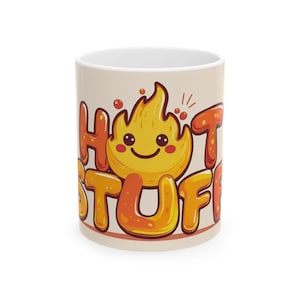 Könnte beinhalten: Keramikbecher mit beigem Hintergrund. Der Becher zeigt die Worte "HOT STUFF" in orangefarbenen und gelben Buchstaben, mit einer lächelnden Flammen-Grafik. Die Innenseite des Bechers ist weiß.