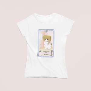 May include: White t-shirt featuring a tarot card design. The card depicts a cartoon cat wearing a cardboard box on its head, labeled "The Hermit." The design is framed in a light blue border with a heart symbol.