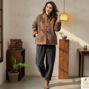 Peut inclure: Une femme porte une veste marron à patchwork avec un col mandarin et des fermetures à bascule, associée à un pantalon gris foncé. La veste présente des carrés aux tons terreux. La tenue est complétée par des bottines marron.